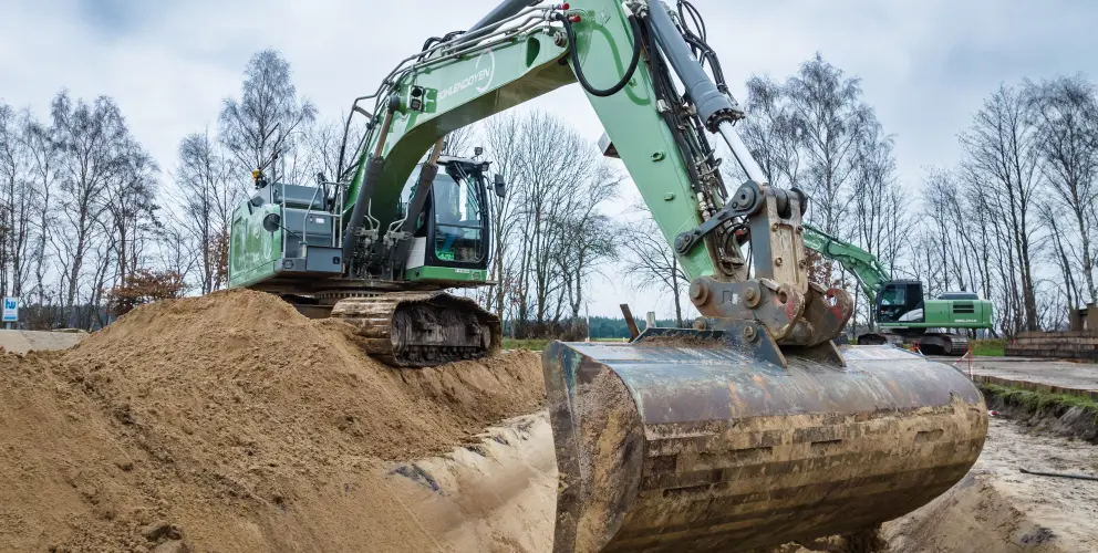Ein großer grüner Bagger bewegt Sand auf einer Baustelle in ländlicher Umgebung, im Hintergrund weitere Baumaschinen und kahle Bäume.