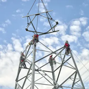 Mehrere Monteure arbeiten gesichert an einem Hochspannungsmast, während ein Kran ein weiteres Mastsegment herablässt – blauer Himmel mit Schäfchenwolken im Hintergrund.