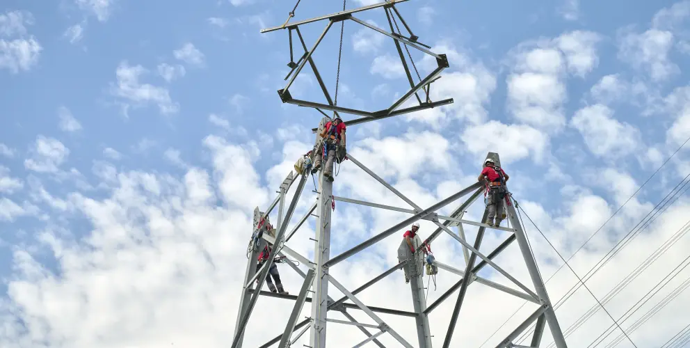 Mehrere Monteure arbeiten gesichert an einem Hochspannungsmast, während ein Kran ein weiteres Mastsegment herablässt – blauer Himmel mit Schäfchenwolken im Hintergrund.