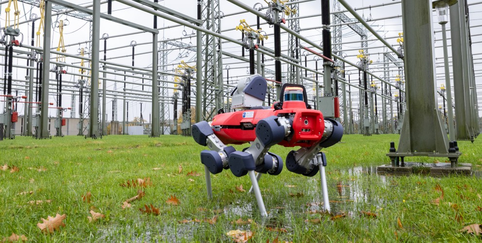 Vierbeiniger Inspektionsroboter mit rotem Gehäuse steht auf nassem Gras in einer Umspannanlage, umgeben von Hochspannungsleitungen und Masten.