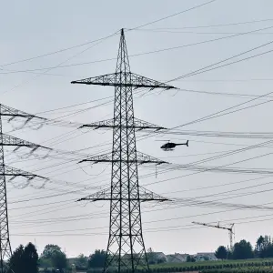 Ein Hubschrauber fliegt nahe an Hochspannungsleitungen zwischen zwei Strommasten entlang – Teil einer Befliegung zur Leitungskontrolle.