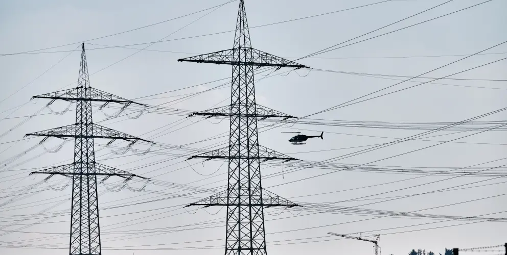 Ein Hubschrauber fliegt nahe an Hochspannungsleitungen zwischen zwei Strommasten entlang – Teil einer Befliegung zur Leitungskontrolle.
