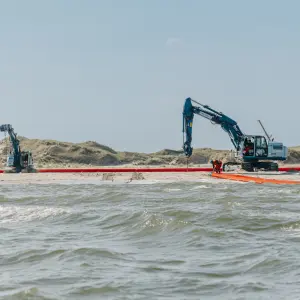Zwei Bagger arbeiten an einem Küstenabschnitt, umgeben von Sanddünen und Wasser, mit mehreren Arbeitspersonen vor Ort.