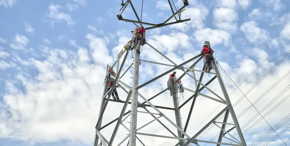Mehrere Monteure arbeiten gesichert an einem Hochspannungsmast, während ein Kran ein weiteres Mastsegment herablässt – blauer Himmel mit Schäfchenwolken im Hintergrund.