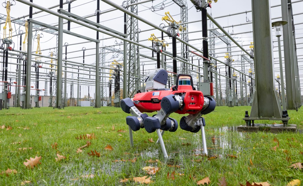 Vierbeiniger Inspektionsroboter mit rotem Gehäuse steht auf nassem Gras in einer Umspannanlage, umgeben von Hochspannungsleitungen und Masten.