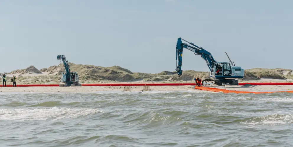 Zwei Bagger arbeiten an einem Küstenabschnitt, umgeben von Sanddünen und Wasser, mit mehreren Arbeitspersonen vor Ort.